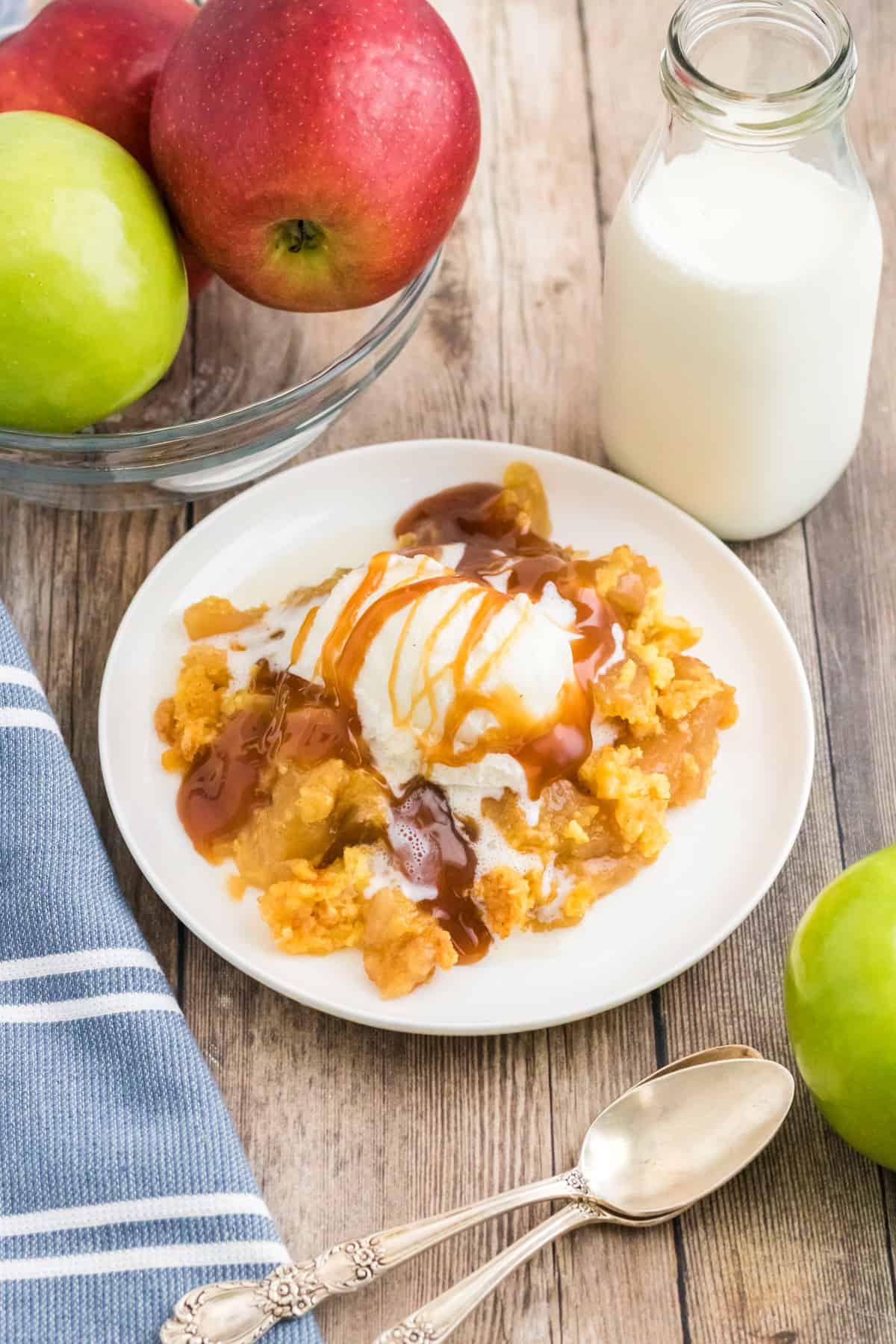 Apple dump cake topped with vanilla ice cream and caramel sauce. Glass of milk and apples are next to plate.