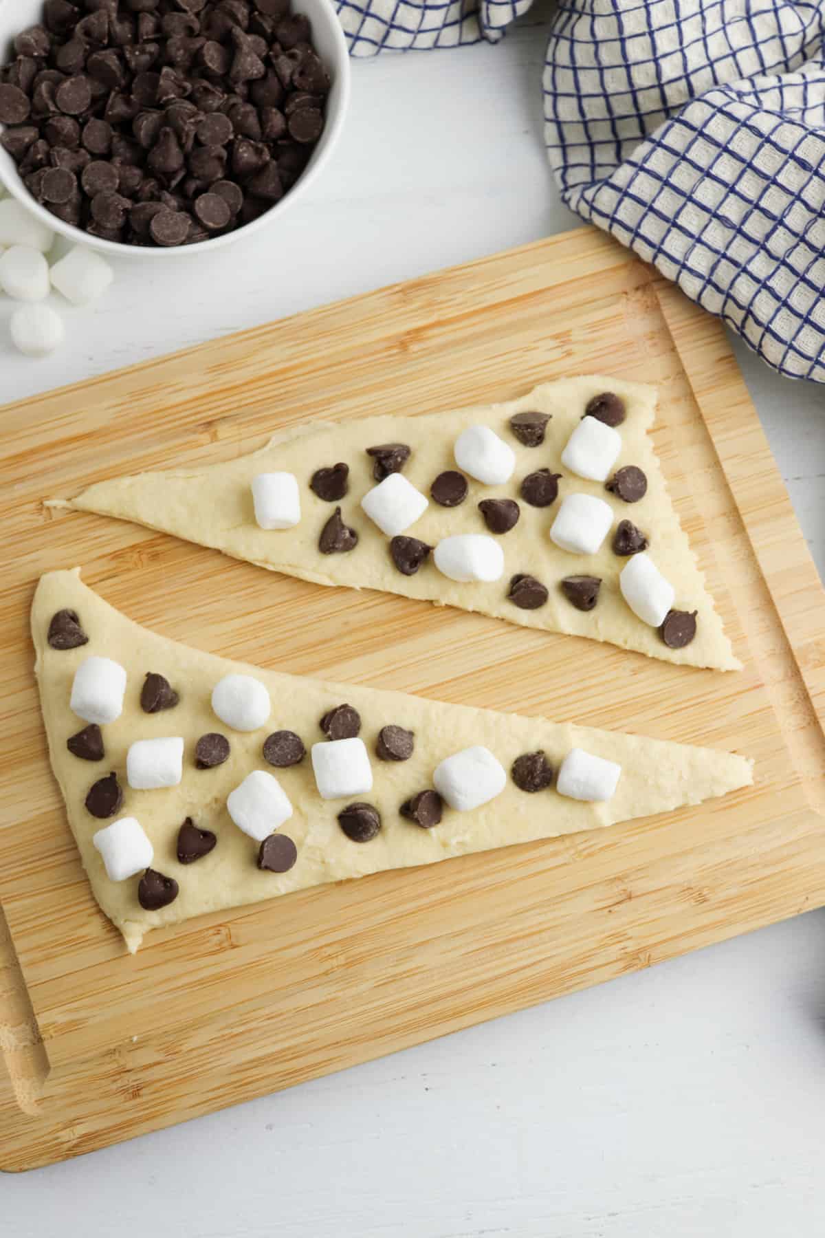 Two triangles of crescent roll dough laid flat on a cutting board and topped with mini marshmallows and chocolate chips.