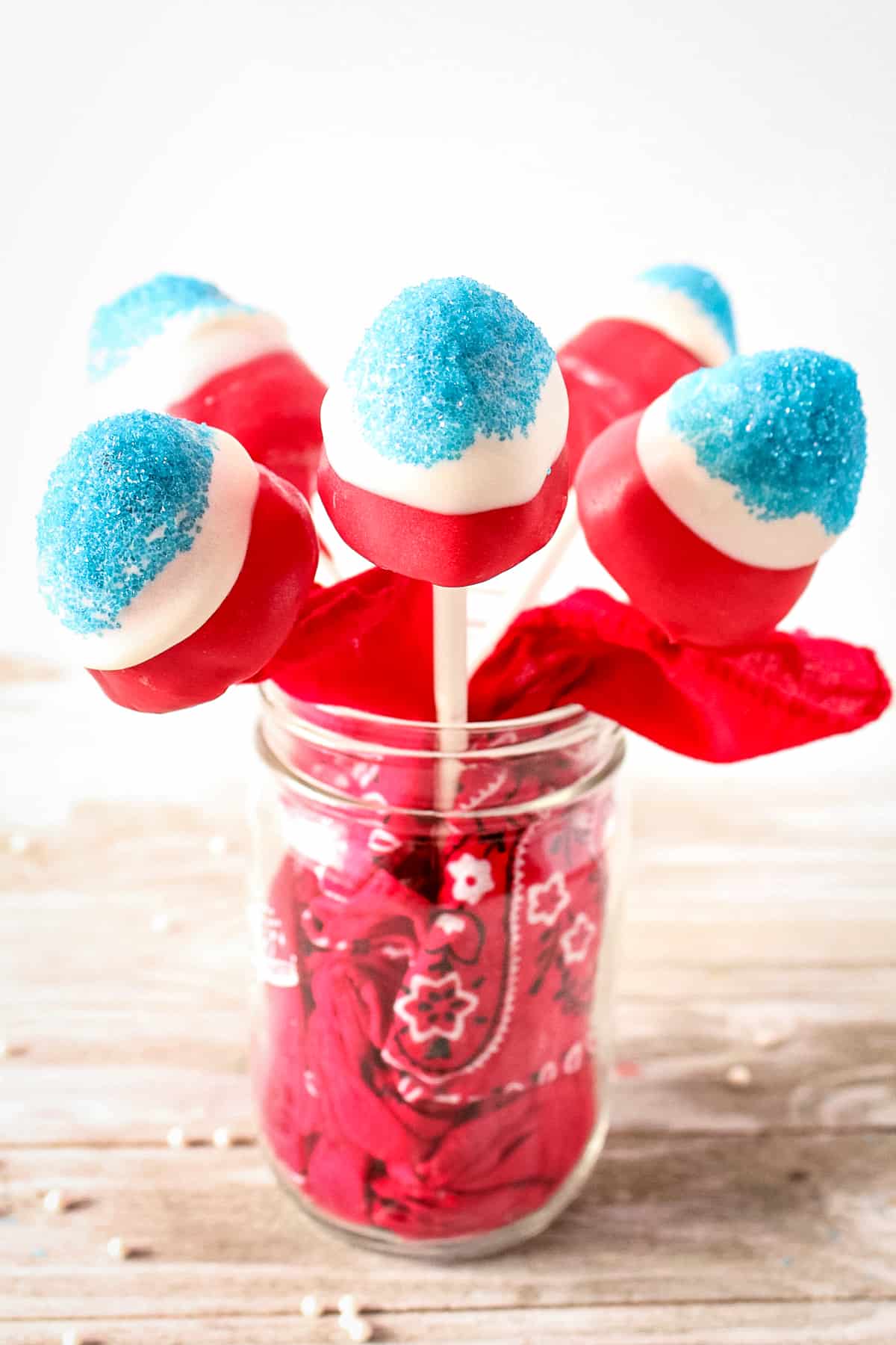 Red, white, and blue covered strawberries on sticks in a mason jar. The mason jar has a red bandana stuffed in it.