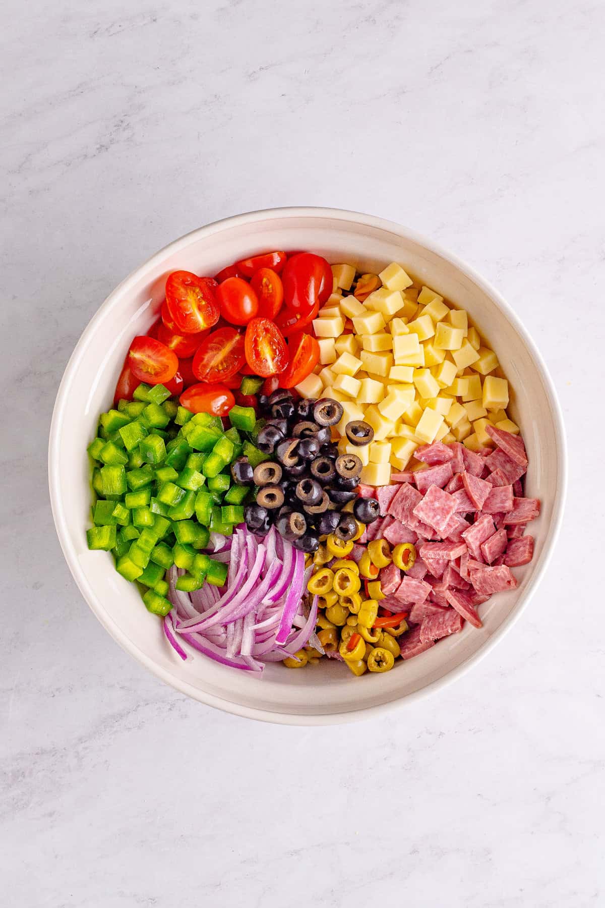 Large bowl with halved grape tomatoes, chopped green pepper, sliced red onion, sliced black and green olives, chopped salami, and cubed mozzarella cheese.