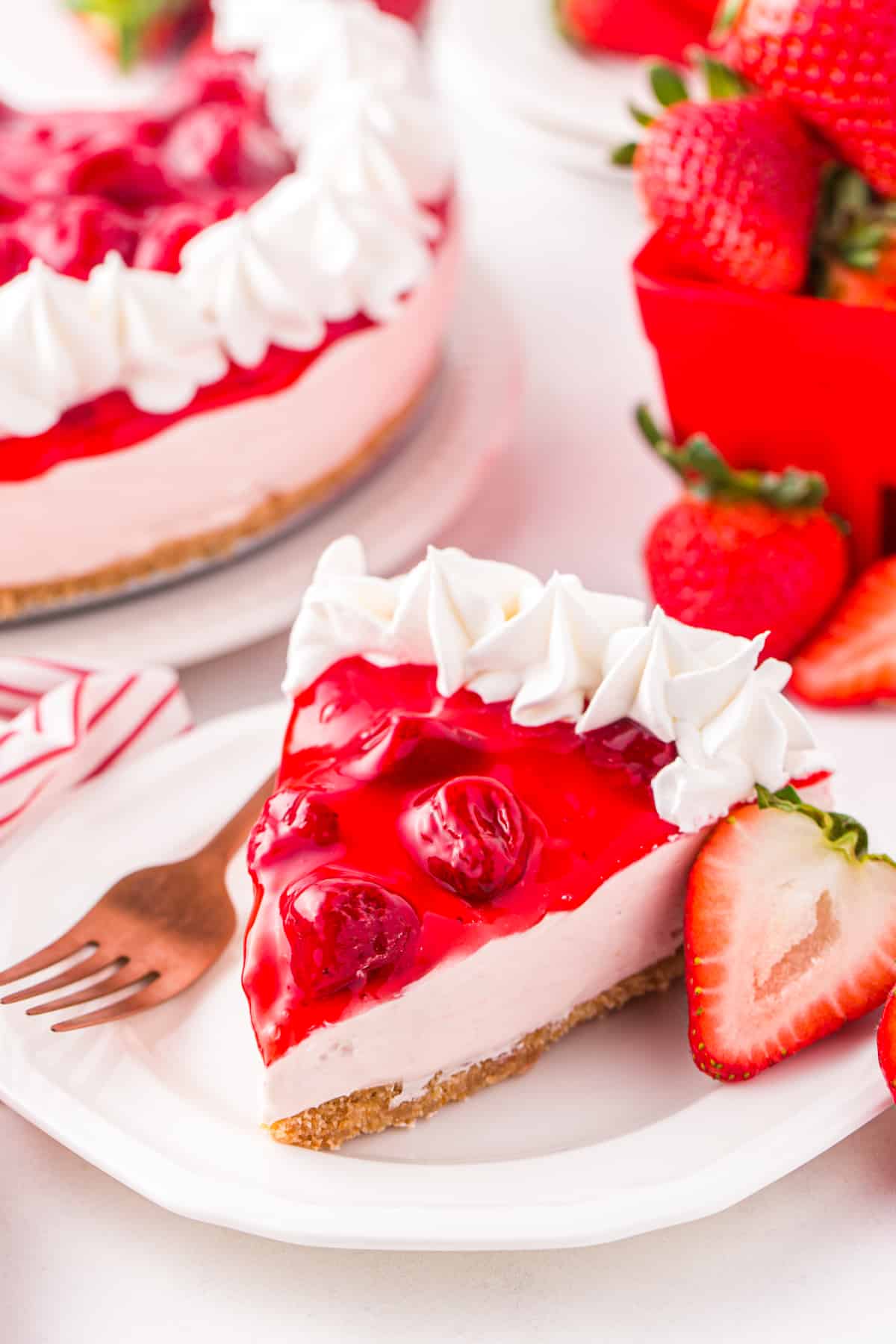 Slice of no bake strawberry cheesecake with strawberry topping and whipped cream piping on edge. Remaining cake and fresh strawberries are in background.