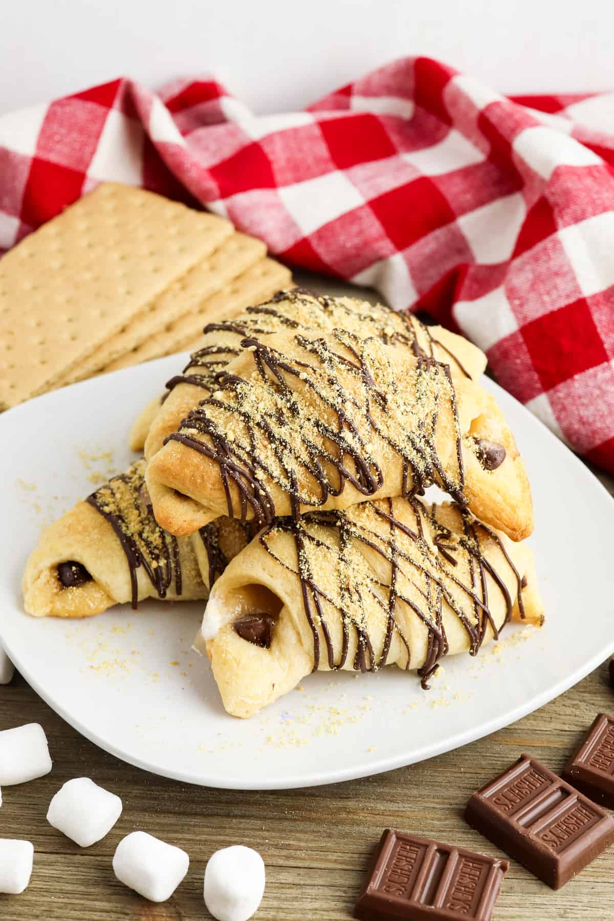 Crescent roll s'mores on white plate with graham crackers, marshmallows, and chocolate around it.
