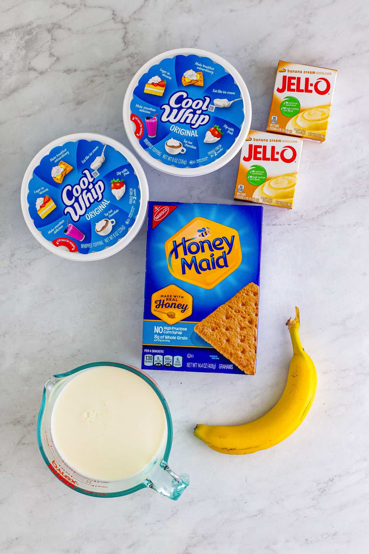 Two containers of Cool Whip, 2 boxes of vanilla instant pudding mix, a box of honey maid graham crackers, a banana, and a measuring cup of milk.