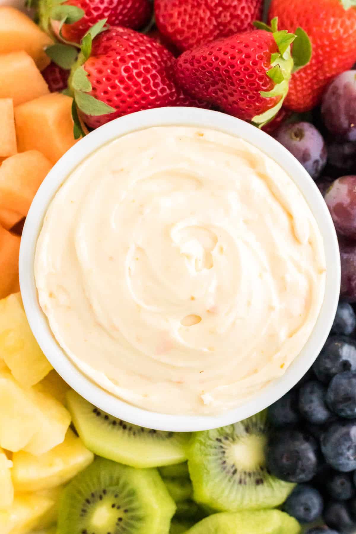 Cream cheese fruit dip in white bowl served with fresh strawberries, pineapple, grapes, kiwi, blueberries, and cantaloupe.