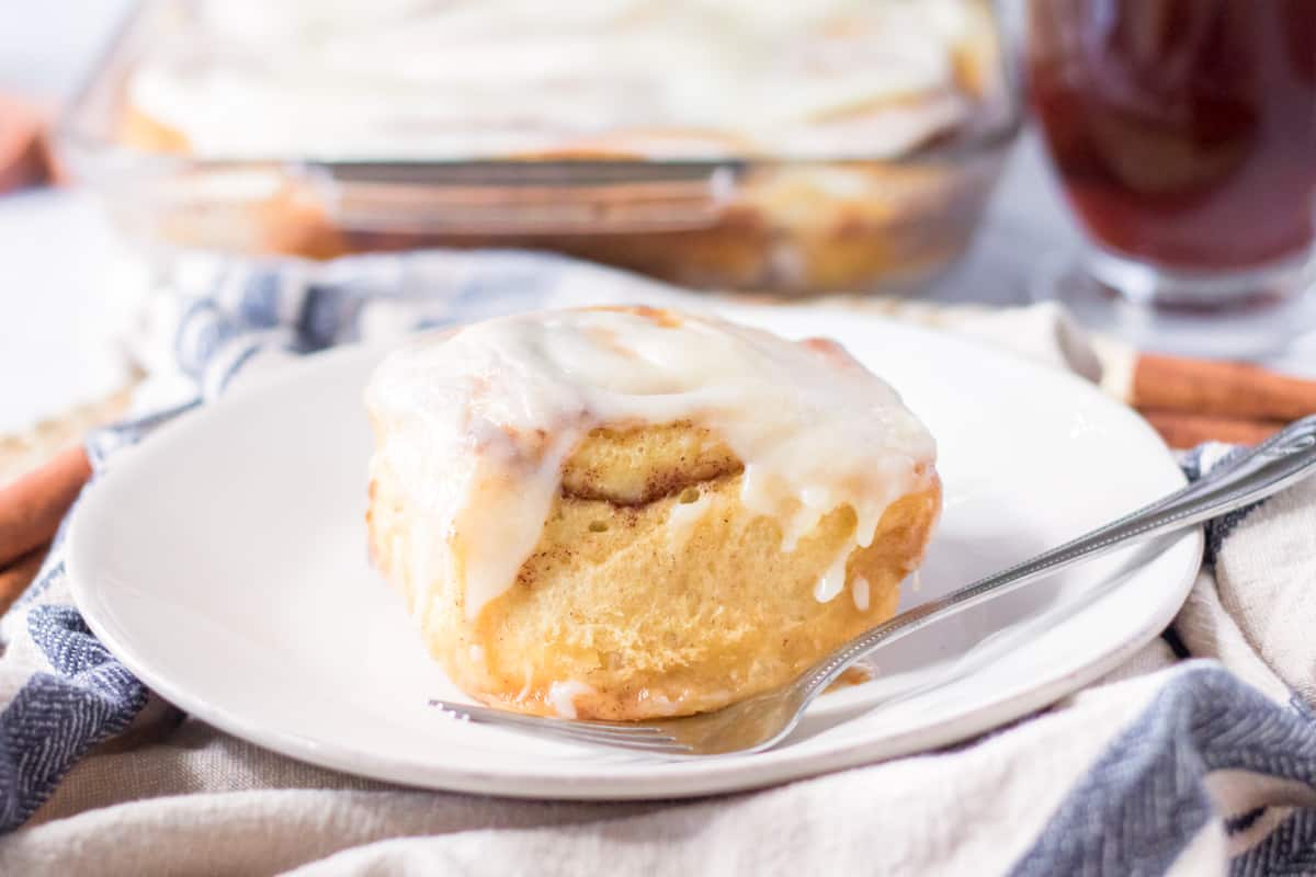 Light and fluffy iced cinnamon roll on plate with dish of rolls behind it.