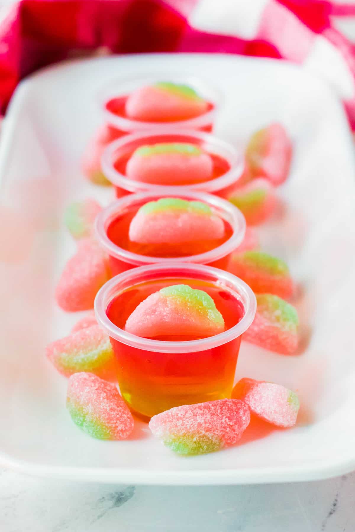 Watermelon vodka jello shots on white serving tray with watermelon sour candies around them.