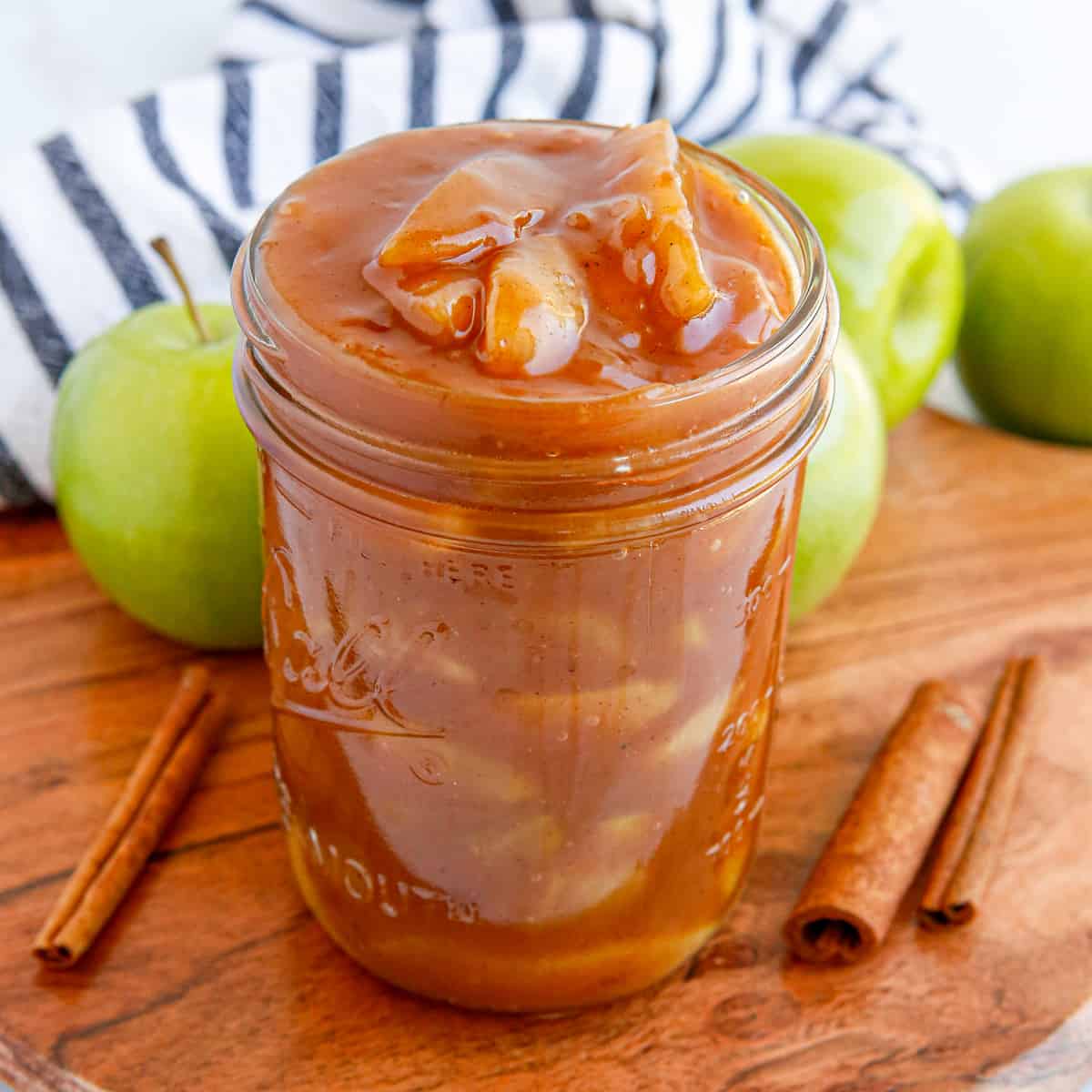 Apple pie filling in a wide mouth mason jar with cinnamon sticks and apples around it.