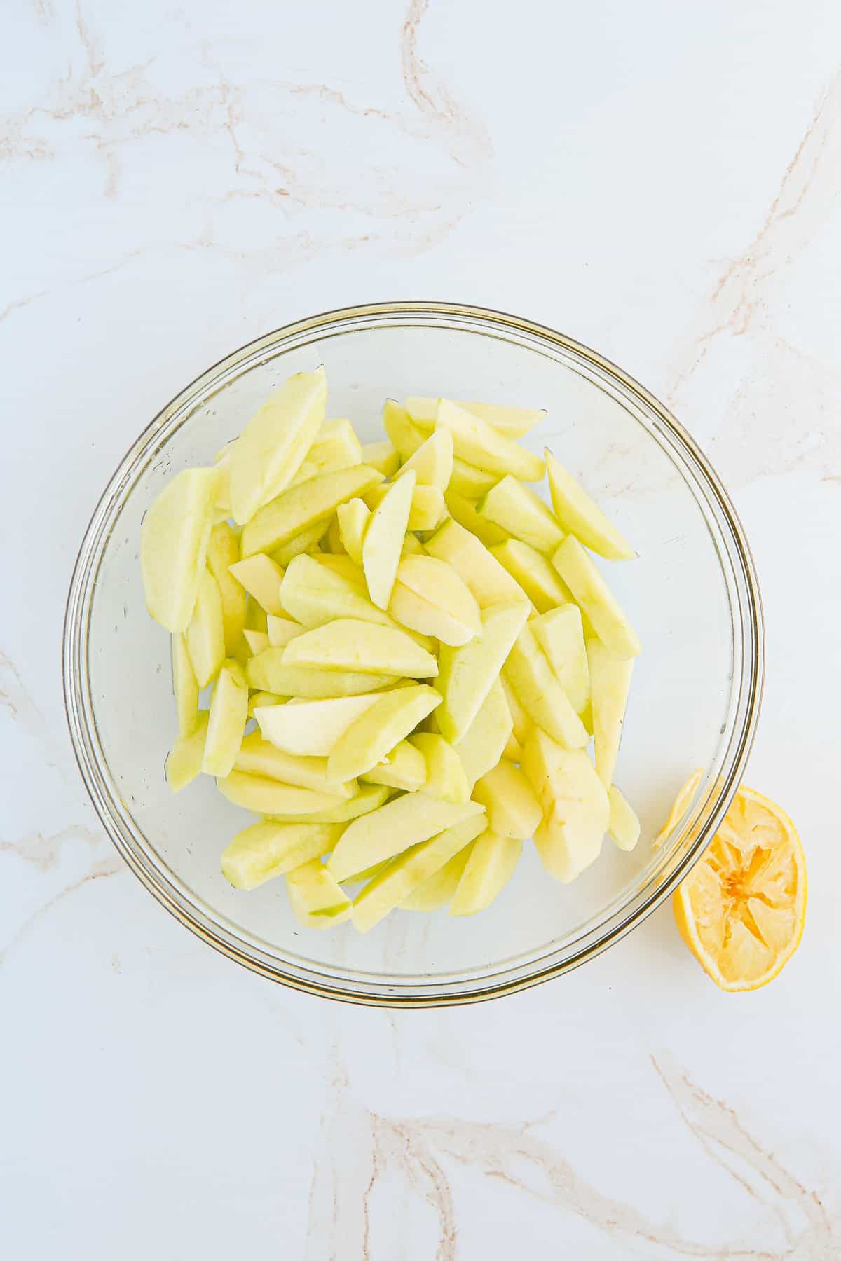 Peeled, cored, and sliced apples in mixing bowl with half a squeezed lemon next to it.