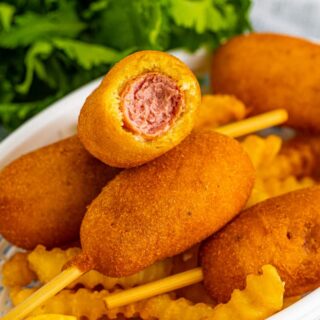 Mini corn dogs in basket with french fries. One corn dog has a bite taken out of it.