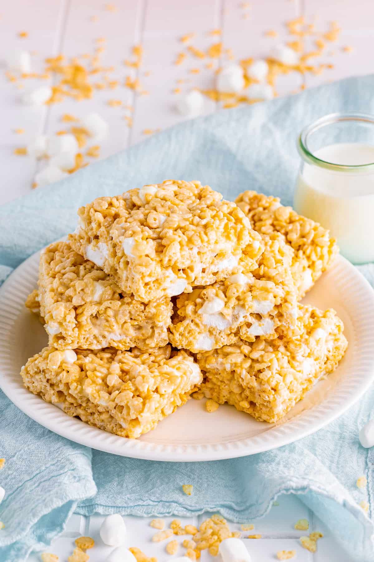 Thick Rice Krispies Treats stacked on white plate with glass of milk in backgroud.