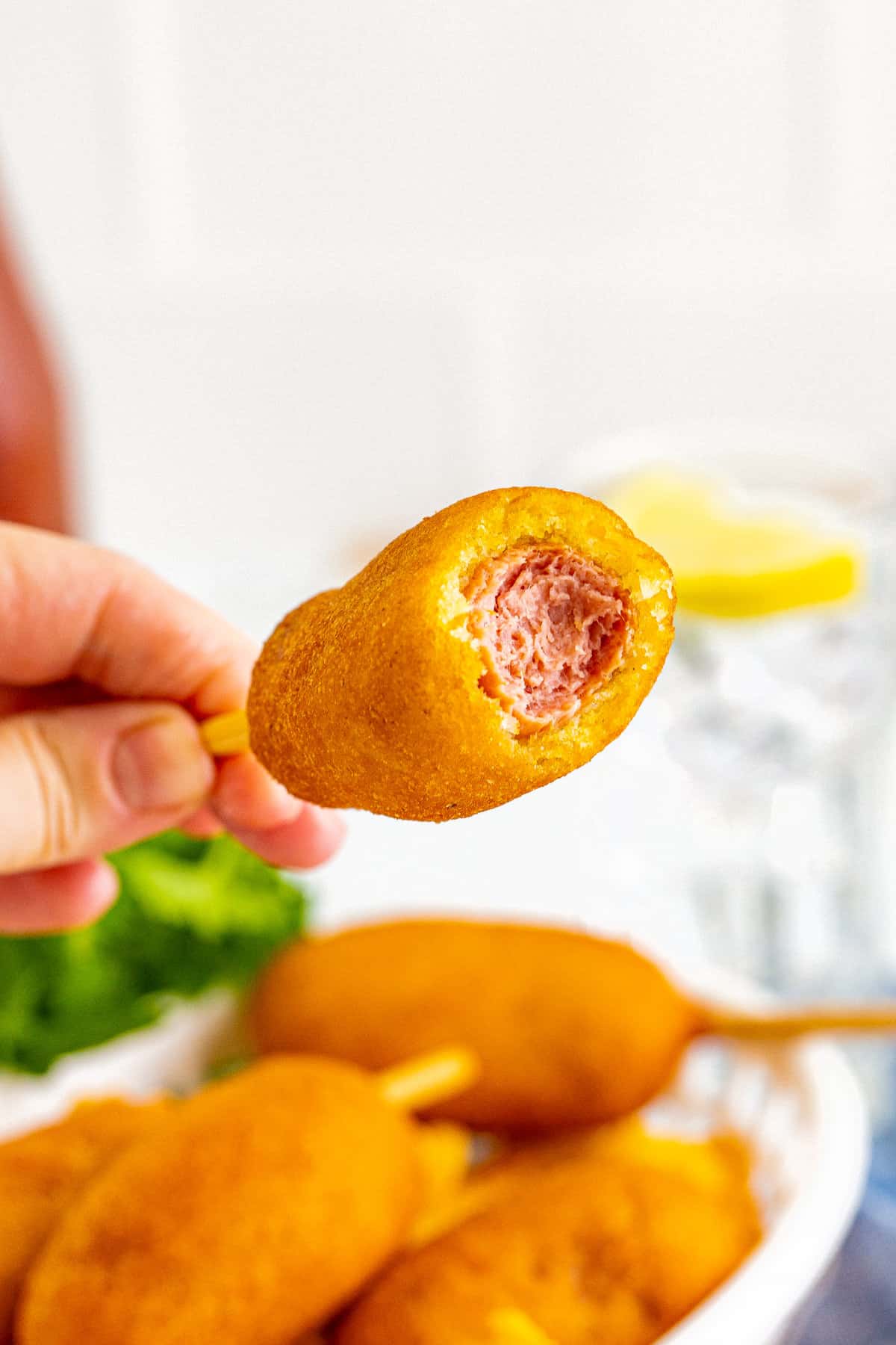 Hand holding mini corn dog with bite taken out of the top to reveal fried dough and hot dog inside.