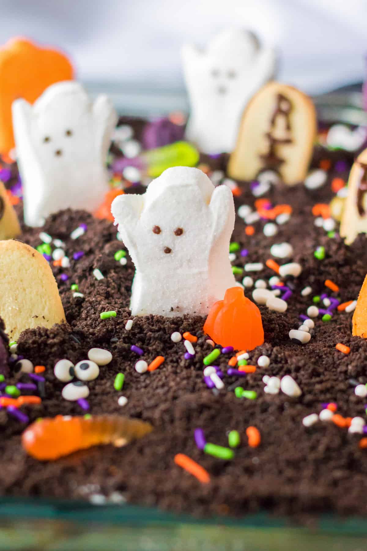 Ghosts in the graveyard halloween poke cake with marshmallow peep ghosts, pumpkins, cats, and cookie tombstones on top.
