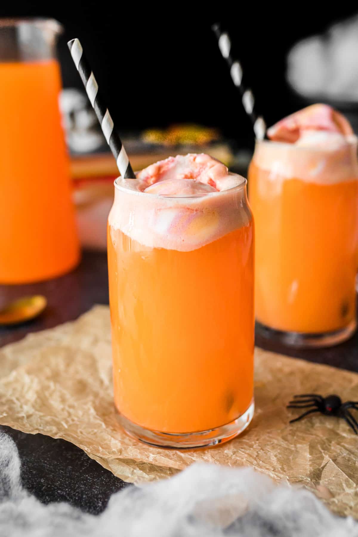 Orange Halloween punch in tall glass topped with sherbet. Another filled glass and pitcher of the punch are in the background.