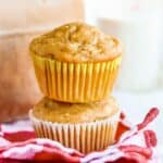 One apple muffin on top of another apple muffin in front of a brown paper bag.