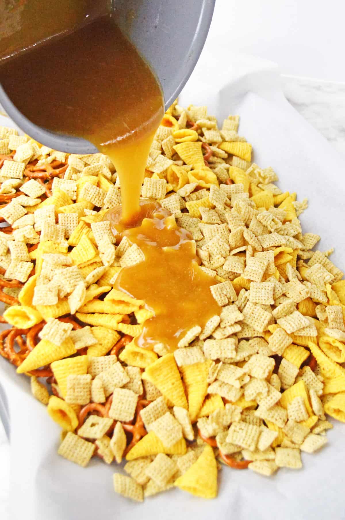 Caramel sauce being poured out of pot and over chex mix on lined baking sheet.