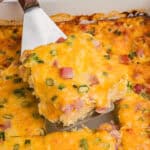 A slice of farmers casserole being lifted from the baking dish, highlighting its chunks of ham, bits of green onion, and golden melted cheese.