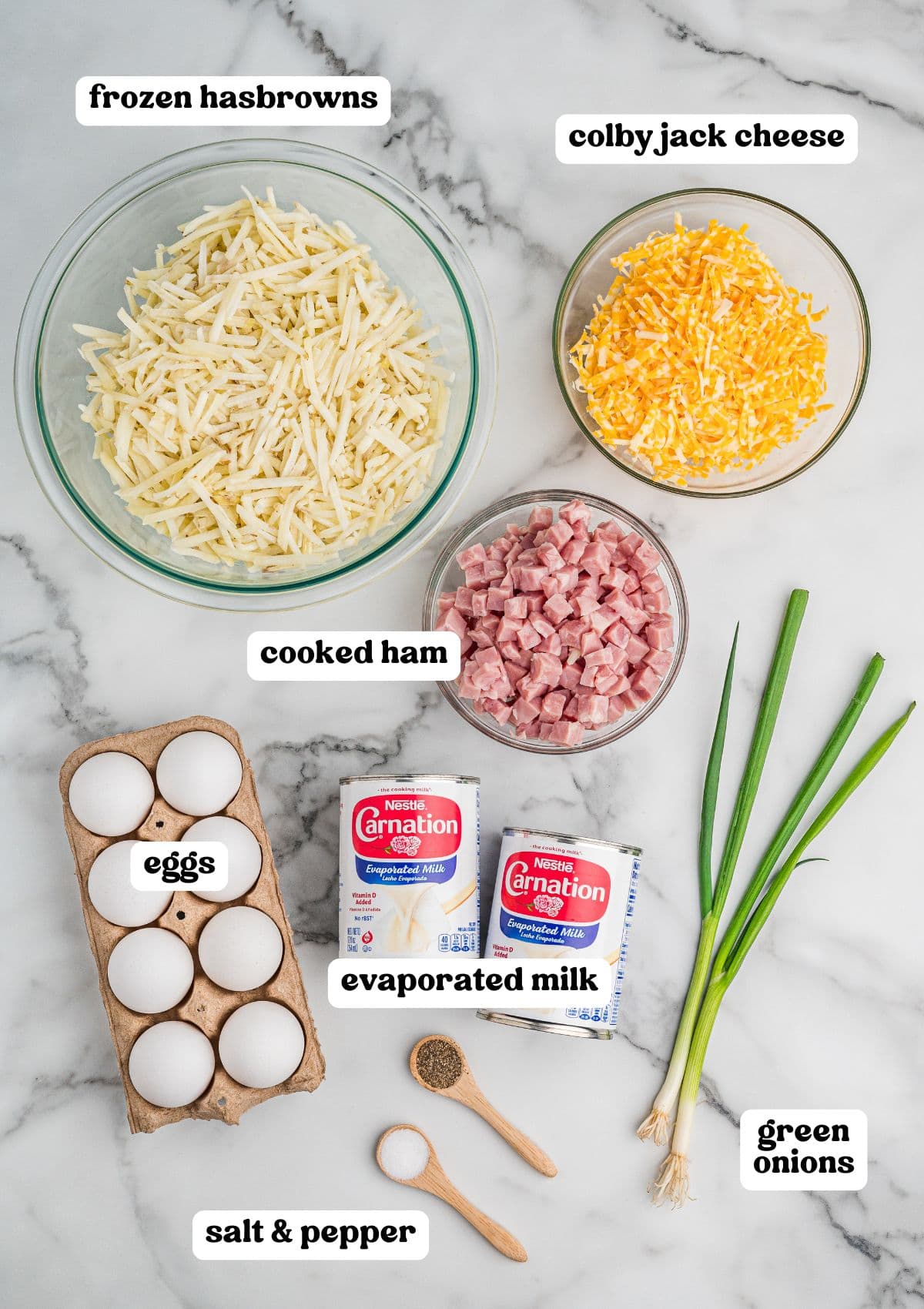 Labeled ingredients on a marble surface, including a bowl of frozen hash browns, shredded Colby Jack cheese, and diced cooked ham, plus eggs, evaporated milk, green onions, and small wooden spoons with salt and pepper.