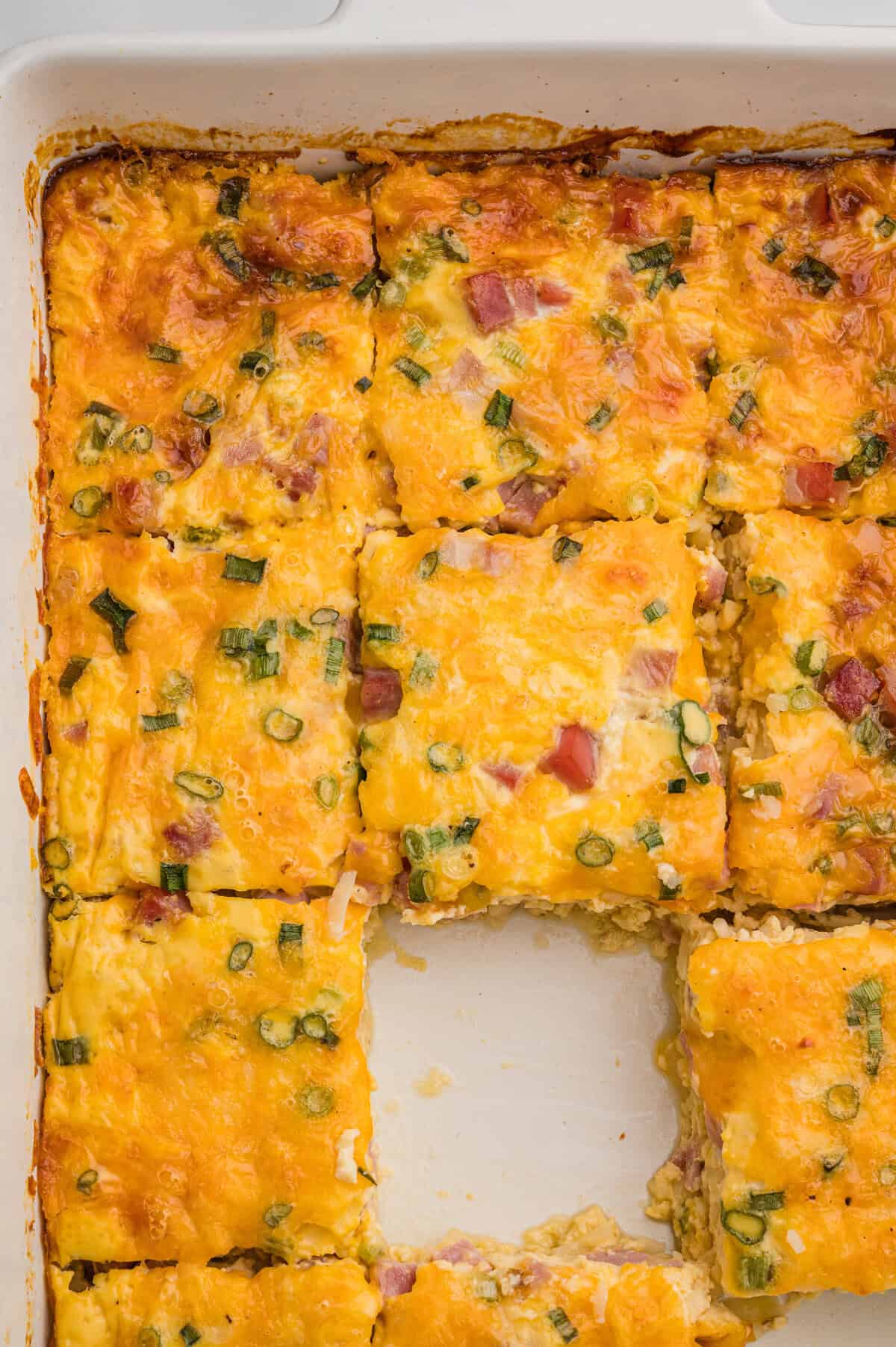 Overhead view of a white baking dish with a sliced breakfast casserole, featuring melted cheese, ham, and green onions, with the center piece removed.