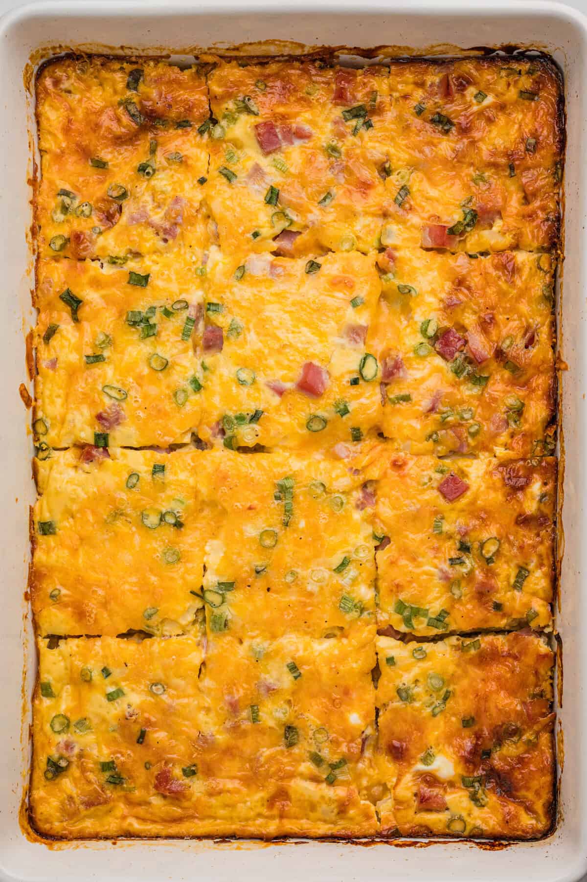 A fully baked farmer's casserole in a white dish, cut into rectangular slices with golden melted cheese, diced ham, and green onion visible on top.