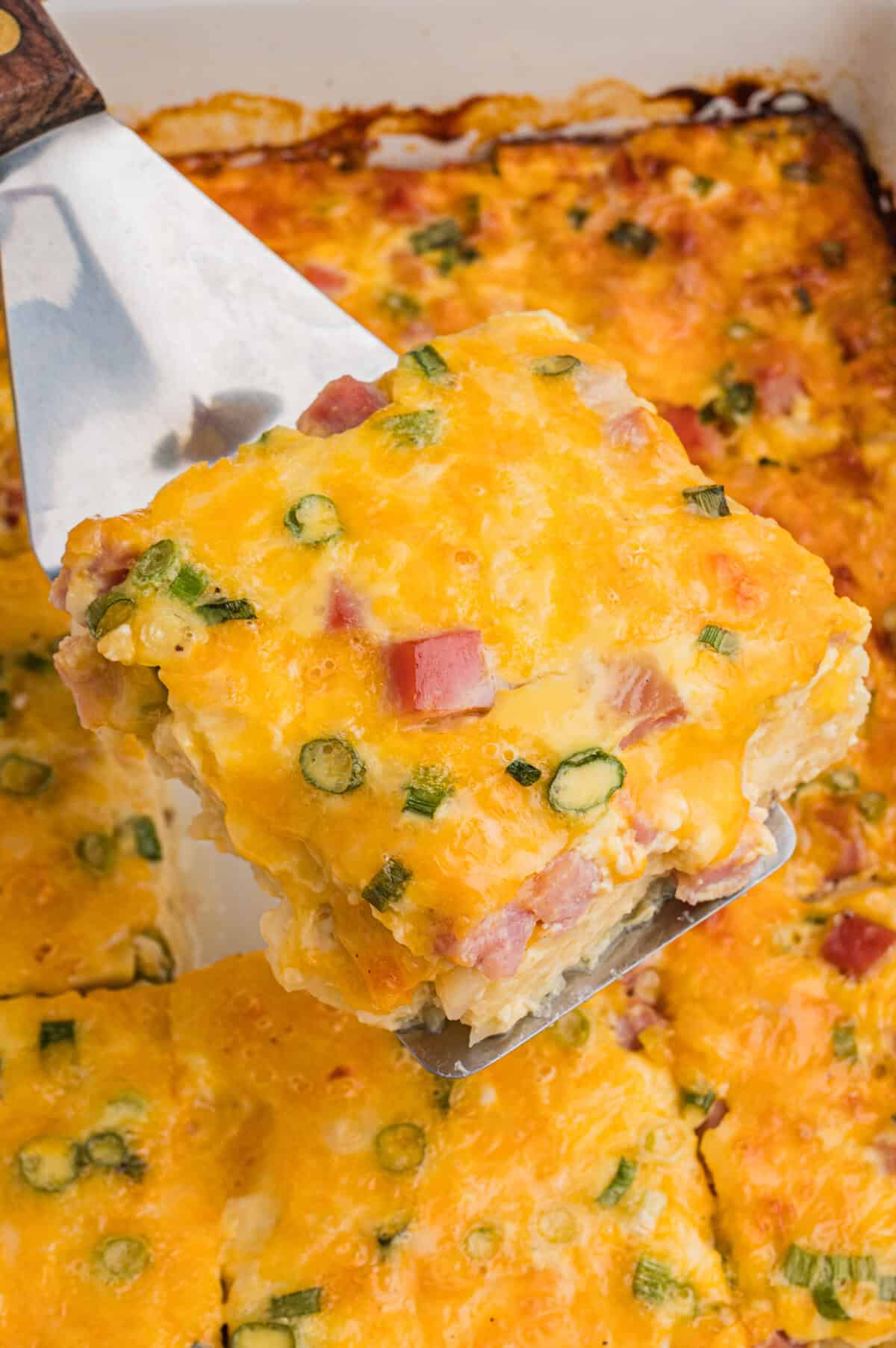 Close-up of a farmers breakfast casserole piece held on a metal spatula, highlighting chunks of ham, bits of green onion, and golden melted cheese.