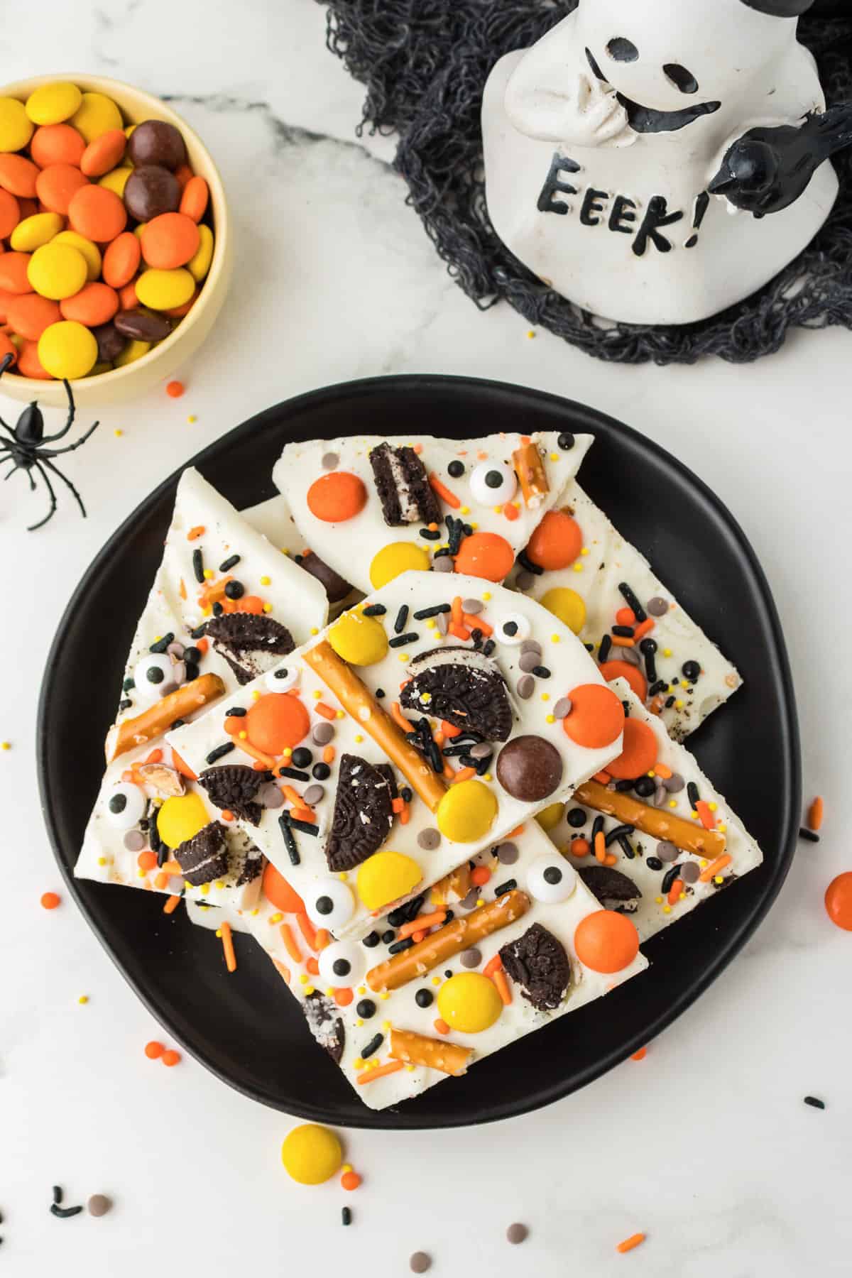 Overhead photo of halloween bark on black plate with bowl of halloween candy and a ghost decoration next to it.
