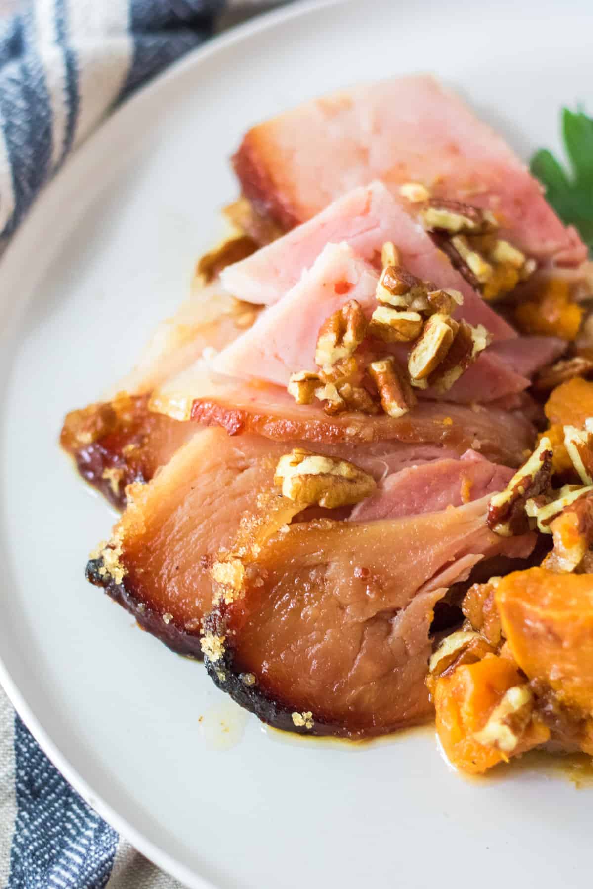 Slices of glazed spiral ham topped with pecans on a white plate with sweet potatoes next to them.