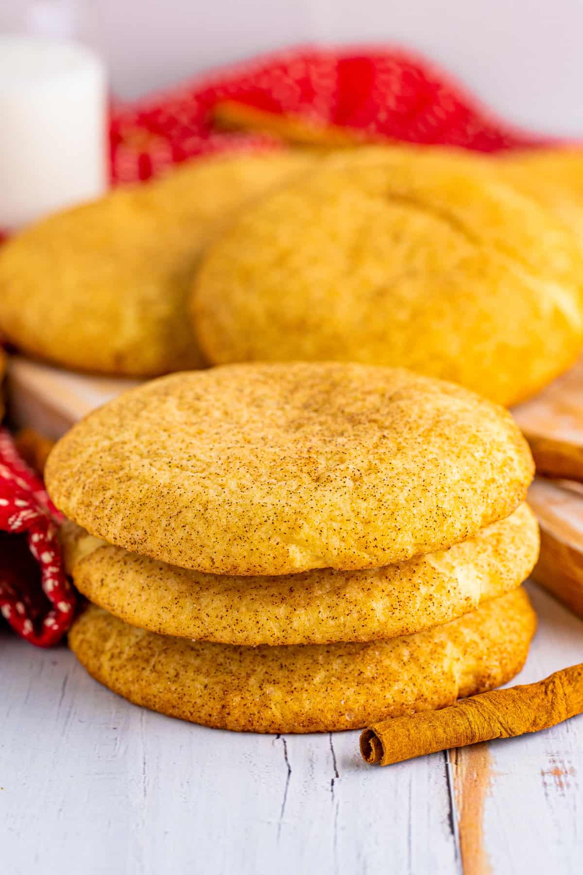 Three large snickerdoodles stacked on tabletop with stick of cinnamon and more cookies around them.