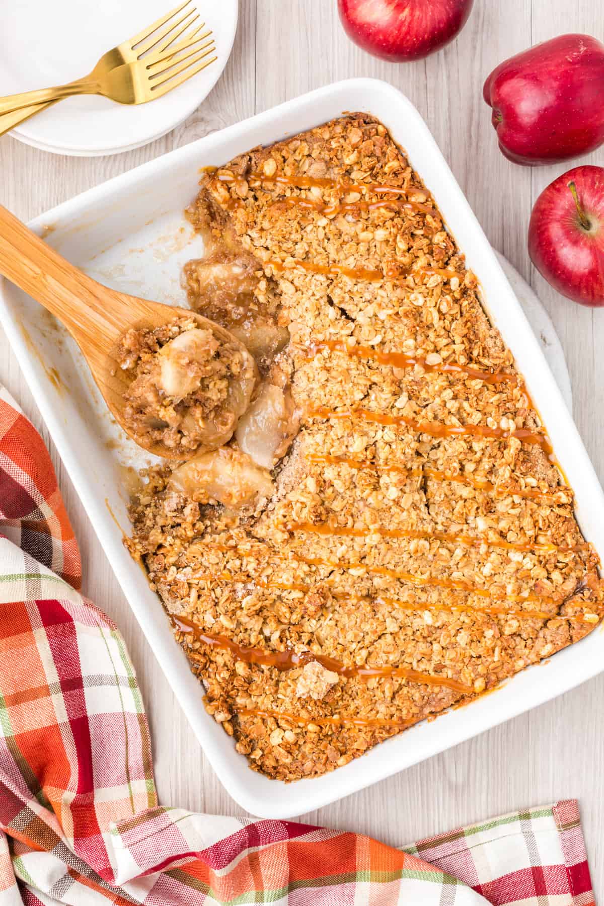 Apple crisp in 9 x 13 baking dish with caramel drizzled on top and a wooded spoon leaning in the removed portion.