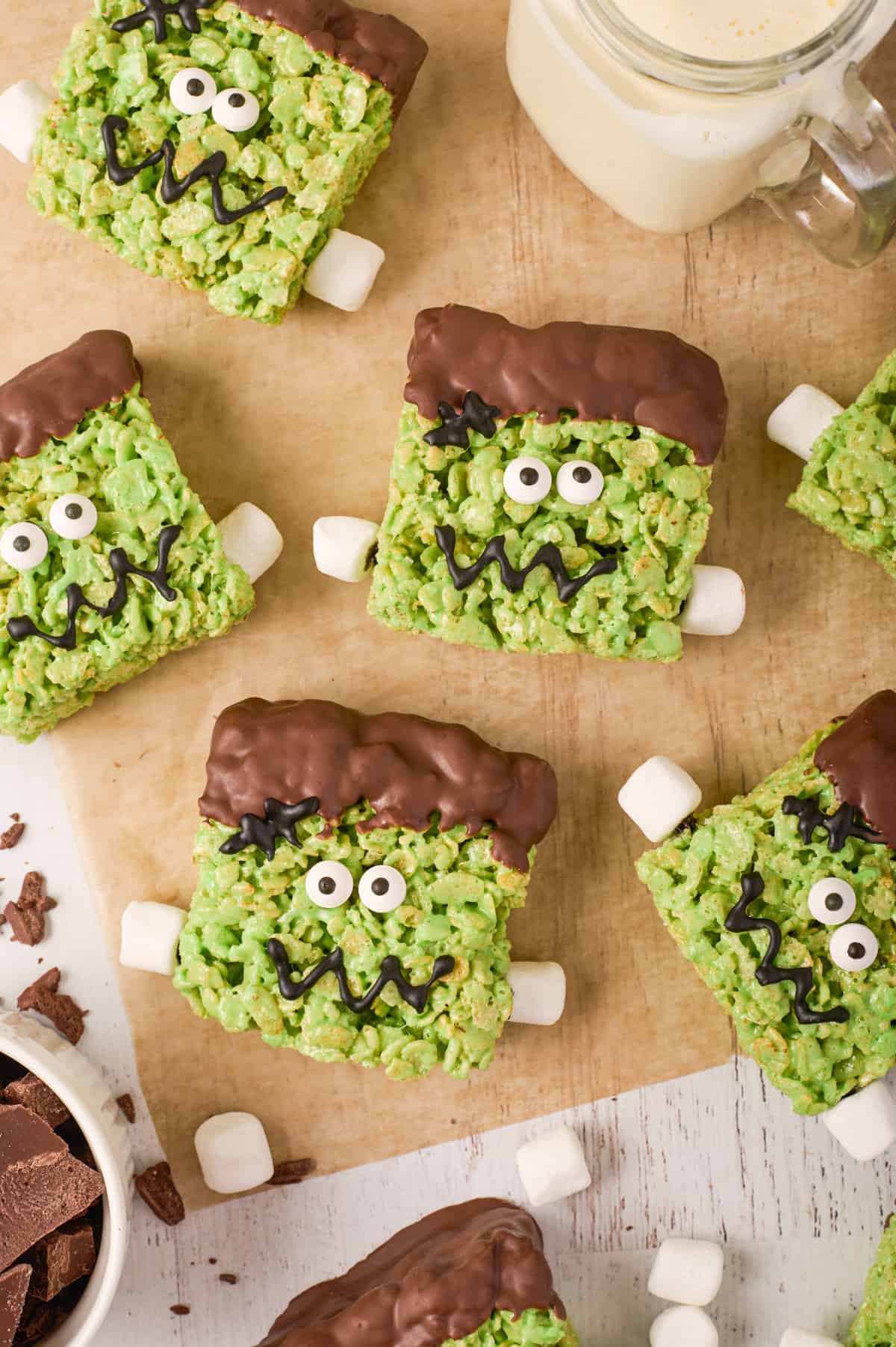 Cute frankenstein rice krispies treats on a piece of parchment paper with mini marshmallows scattered and a glass of milk.