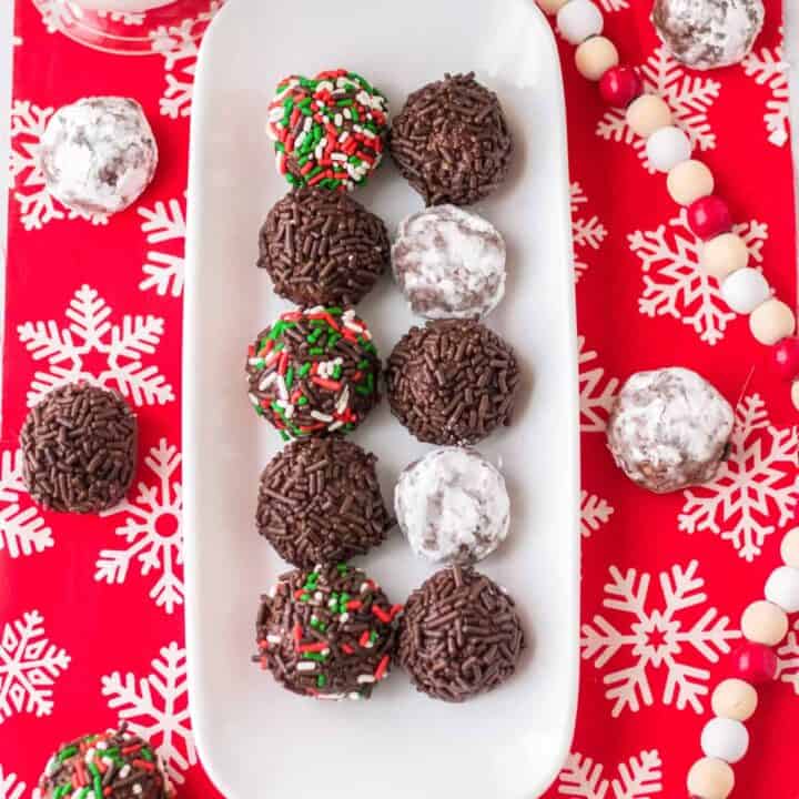 Chocolate rum balls coated in powdered sugar or sprinkles and on a white serving tray over a holiday table cloth.