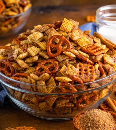 Cinnamon sugar chex mix in glass serving bowl with measuring cup of brown sugar and cinnamon sticks beside it.d