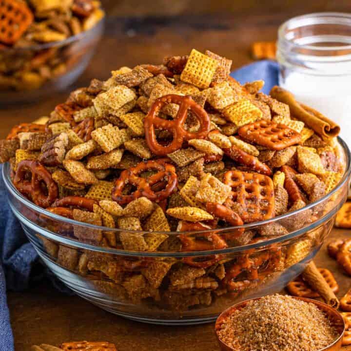 Cinnamon sugar chex mix in glass serving bowl with measuring cup of brown sugar and cinnamon sticks beside it.d