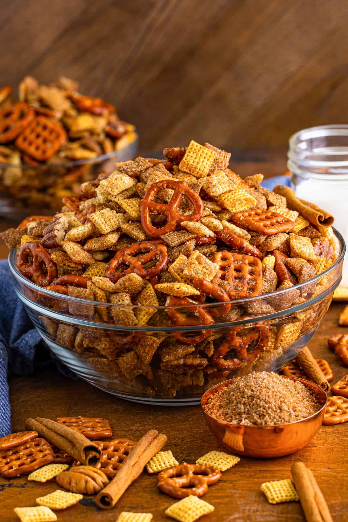 Cinnamon sugar chex mix in glass serving bowl with measuring cup of brown sugar and cinnamon sticks beside it.