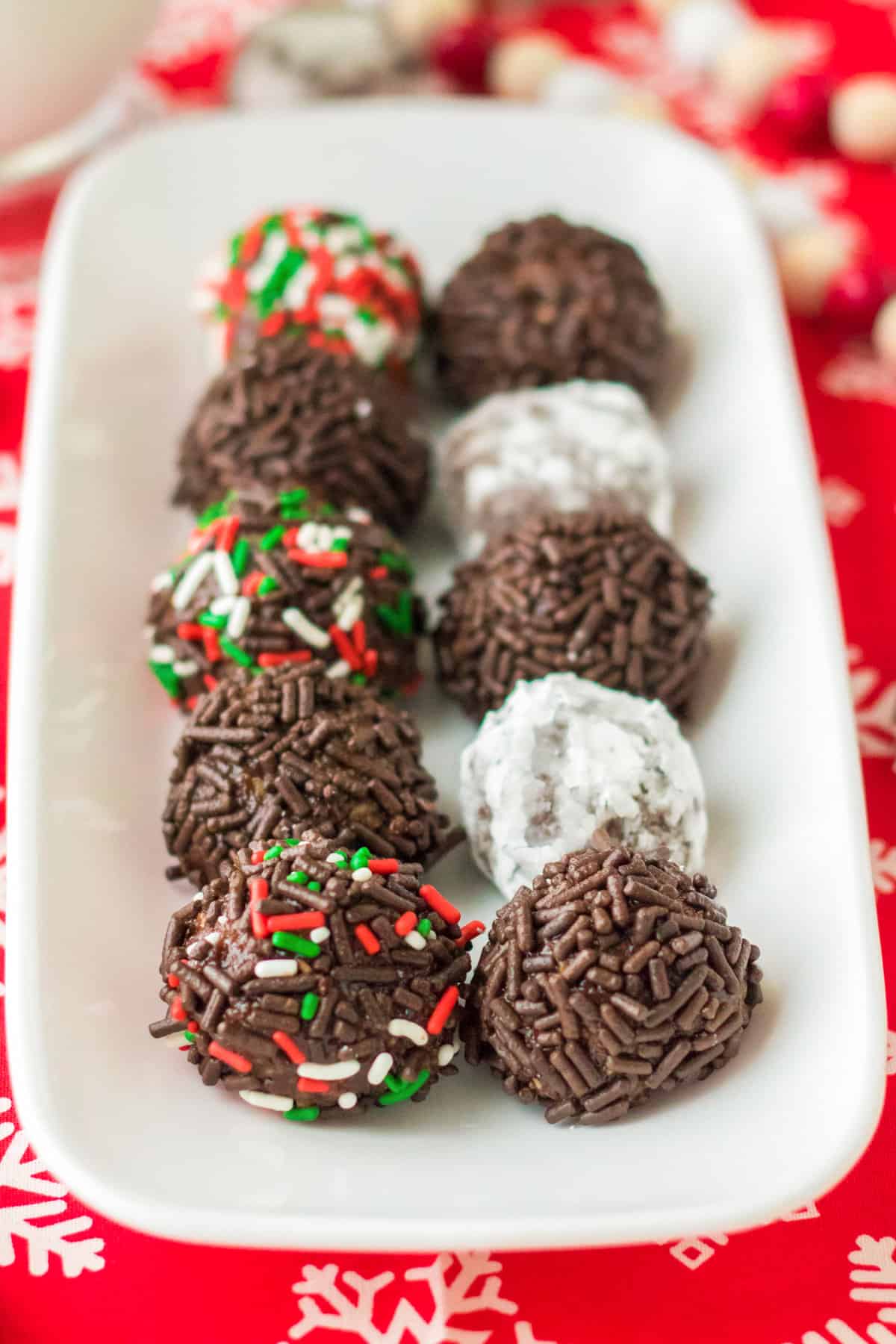 No bake chocolate rum balls on a white serving tray. Some are coated in powdered sugar others in sprinkles.