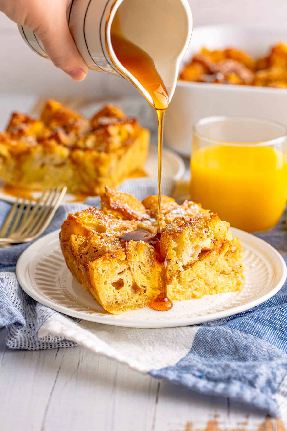 Slice of French toast casserole on white plate with maple syrup being poured over the top. Glass of orange juice and another piece of the bake are in background.
