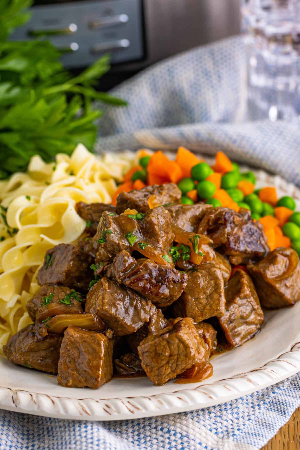 Steak bites, noodles, and vegetables on white plate with glass of water and slow cooker in background.