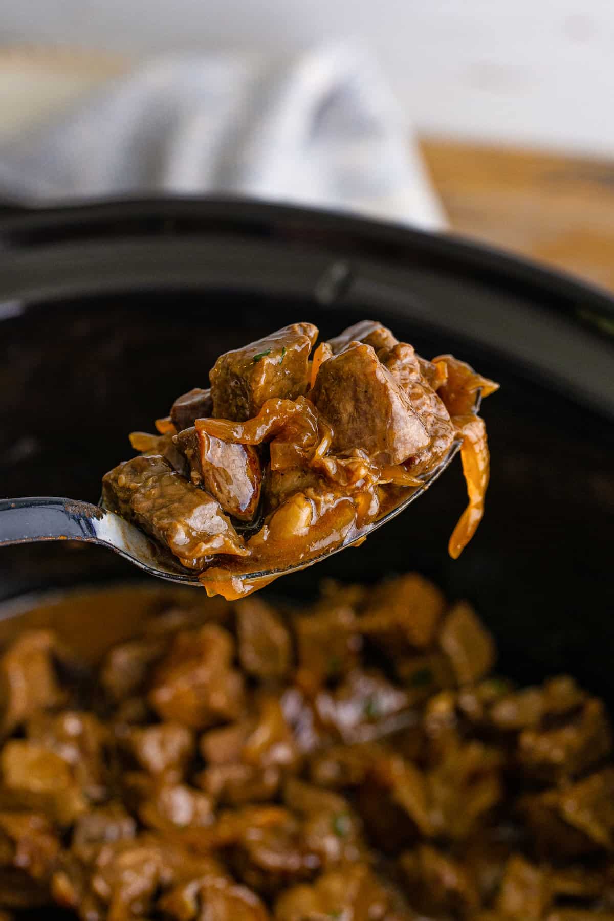 Serving spoon removing a spoonful of steak bites with onions from the slow cooker.