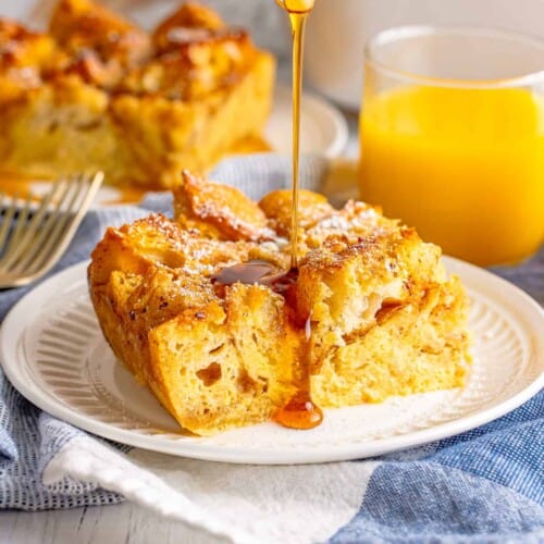 Slice of French toast casserole on white plate with maple syrup being poured over the top. Glass of orange juice and another piece of the bake are in background.