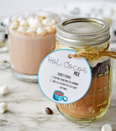 Homemade hot cocoa mix in a mason jar with a gift tag and a mug of hot chocolate with marshmallows behind it.