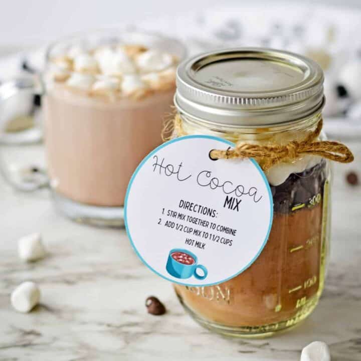 Homemade hot cocoa mix in a mason jar with a gift tag and a mug of hot chocolate with marshmallows behind it.