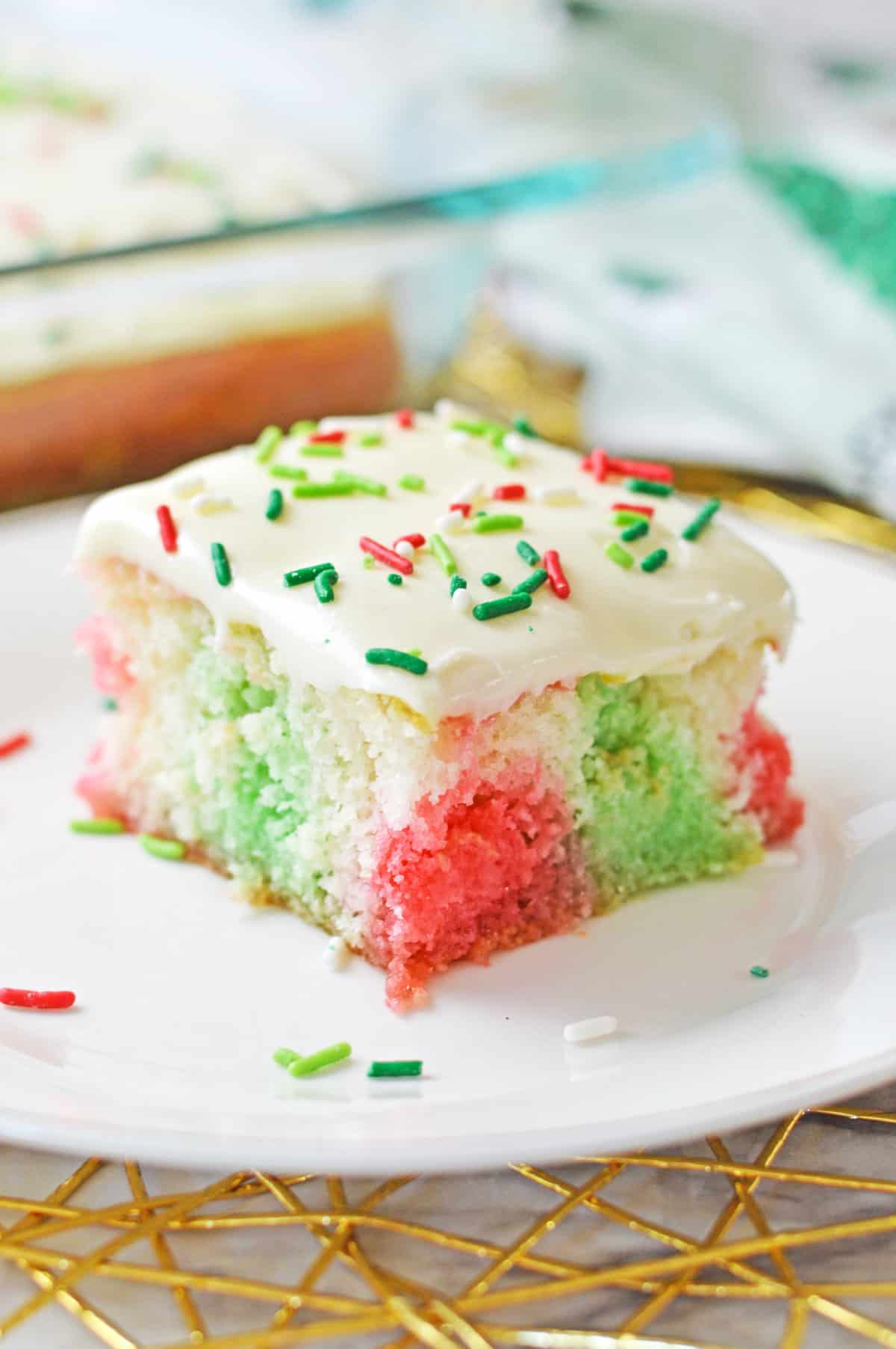 Christmas poke cake with pockets of red and green color throughout a fluffy white sheet cake and vanilla frosting with red and green sprinkles.