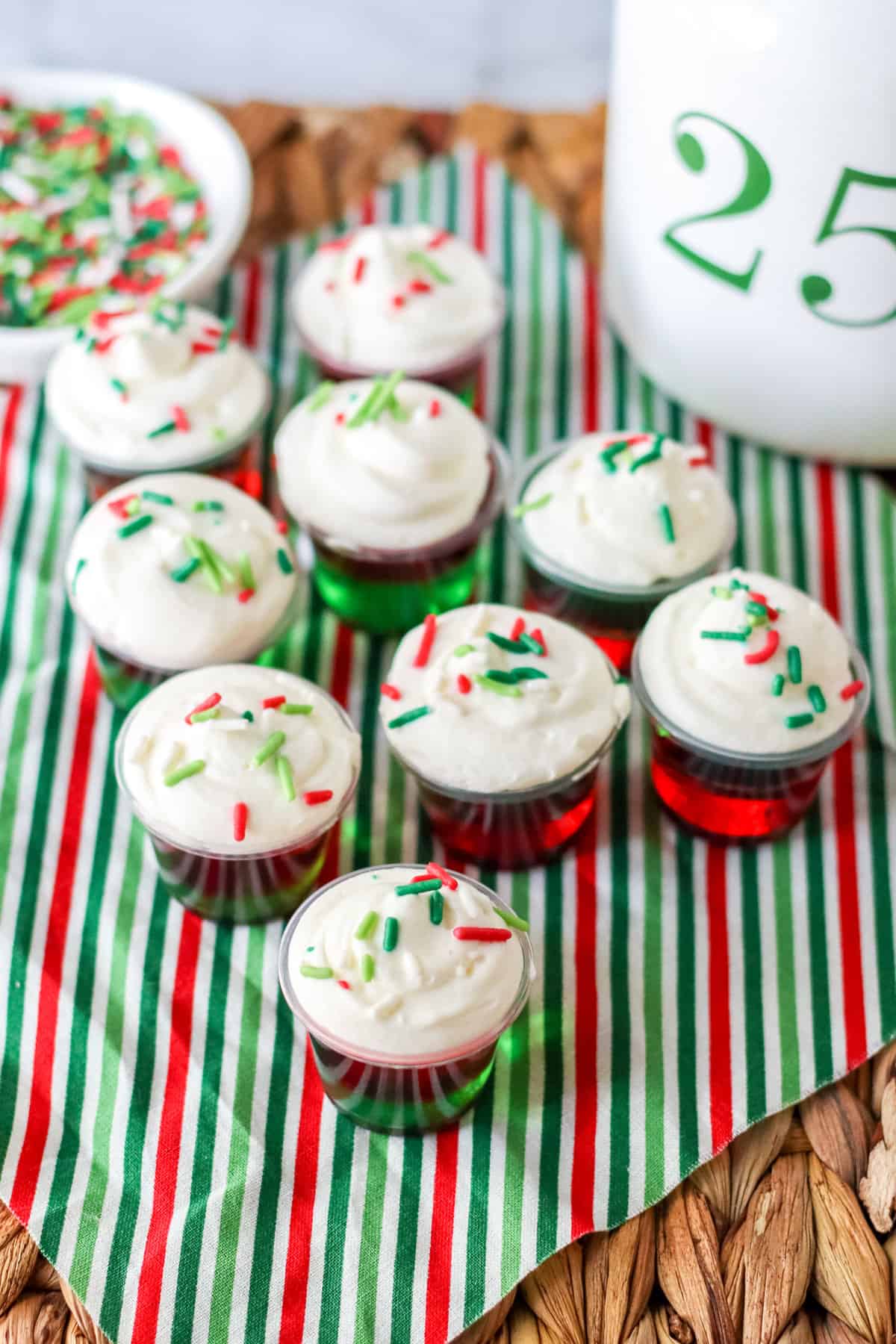Several Christmas jello shots placed on festive holiday napkin with bowl of red and green sprinkles in the background.