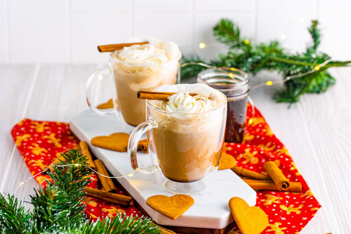 Gingerbread lattes on marble tray topped with whipped cream and cinnamon sticks and surrounded by holiday decor.