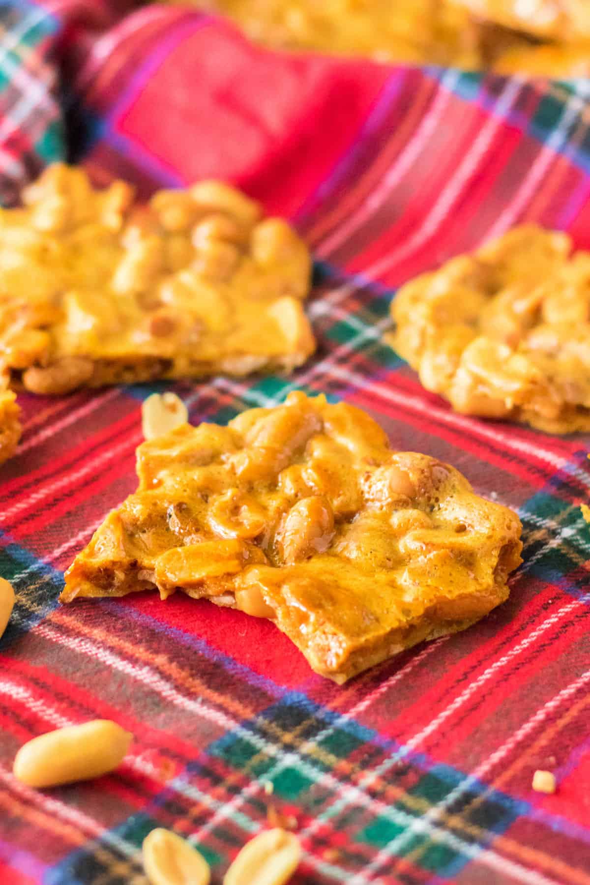 Pieces of light and crunchy peanut brittle on red plaid tablecloth with roasted peanuts next to them.