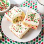 Thick Christmas sugar cookie bars with sprinkles in the dough and topped with vanilla frosting and more sprinkles. A glass of milk and bowl of holiday chips can be seen around them.