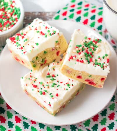 Thick Christmas sugar cookie bars with sprinkles in the dough and topped with vanilla frosting and more sprinkles. A glass of milk and bowl of holiday chips can be seen around them.