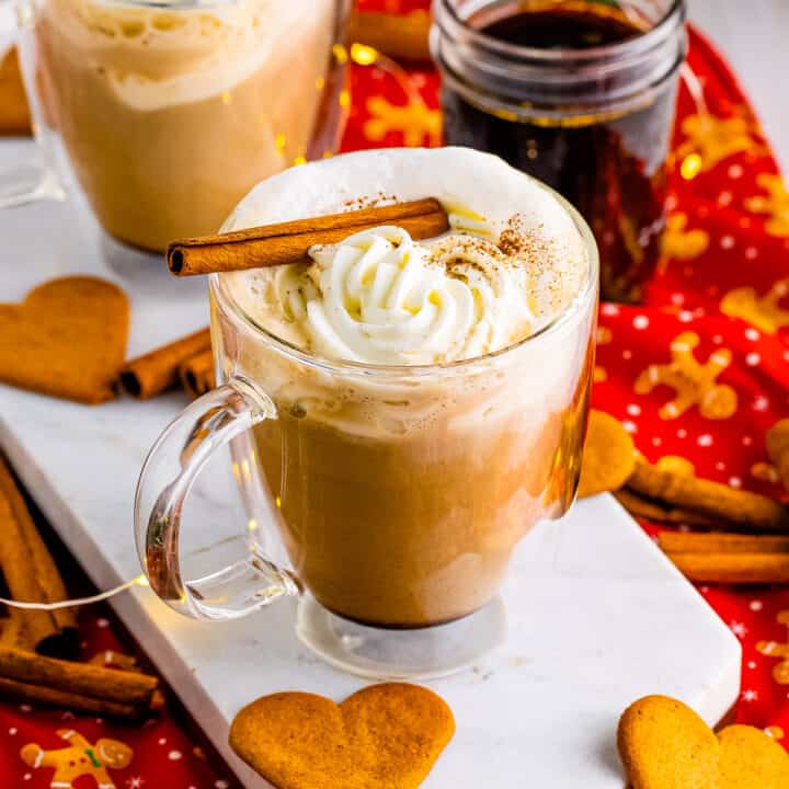 Homemade gingerbread latte in glass mug topped with whipped cream and a cinnamon stick. Heart-shaped gingerbread cookies are next to the hot drink.