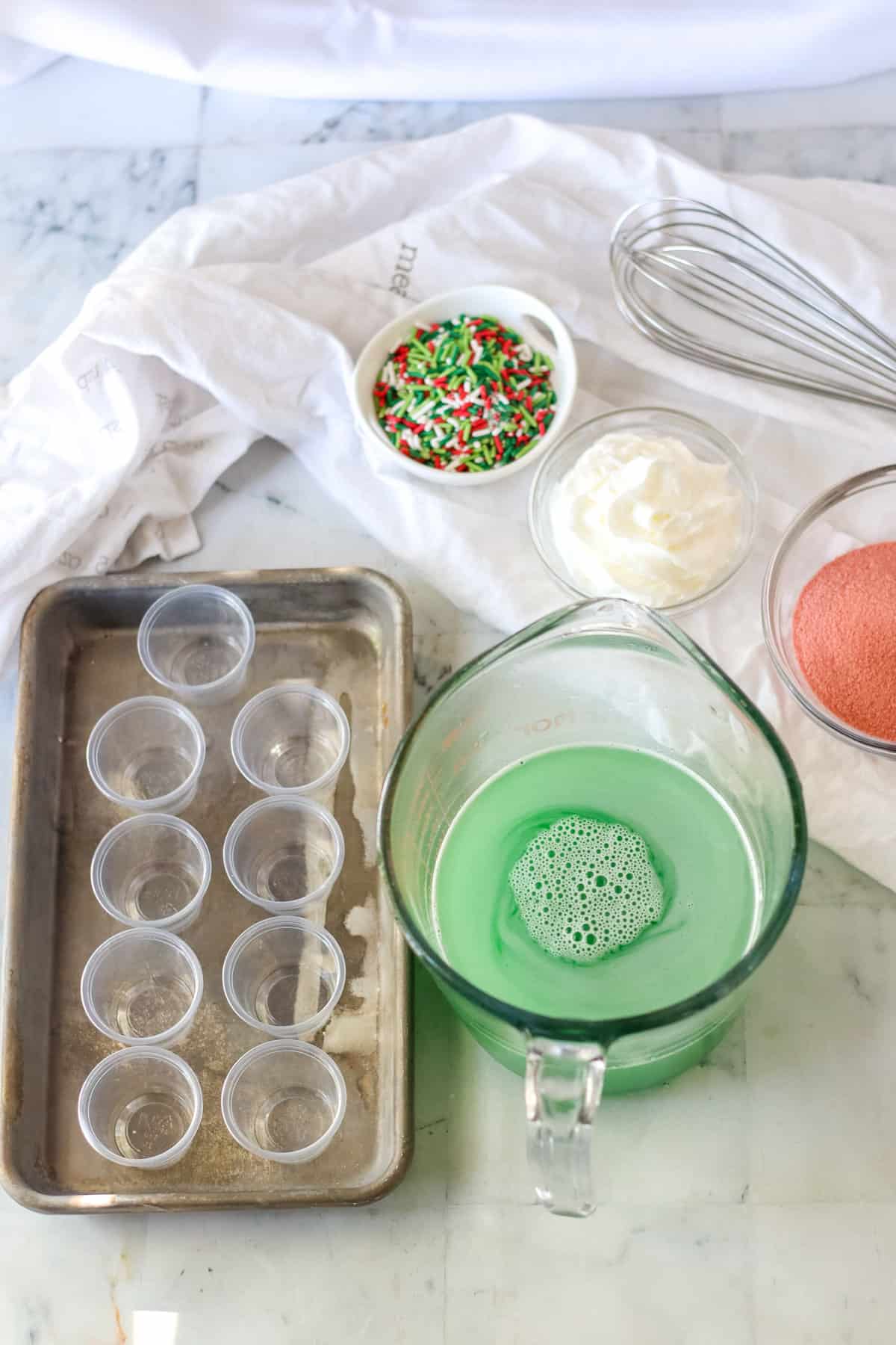 Large measuring cup with green jello mixture next to tray of shot glasses.