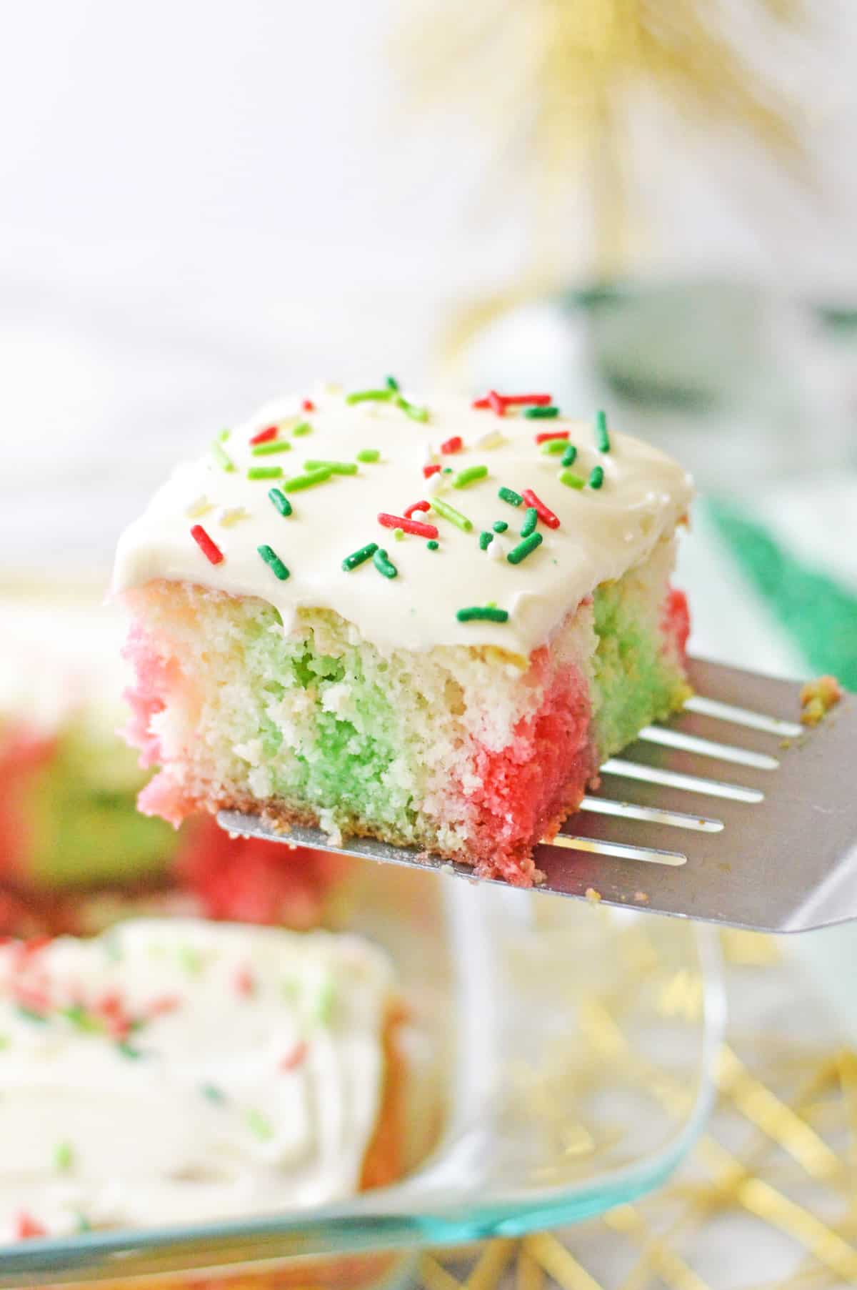 Square slice of red and green jello poke cake being lifted out of baking dish with metal spatula.