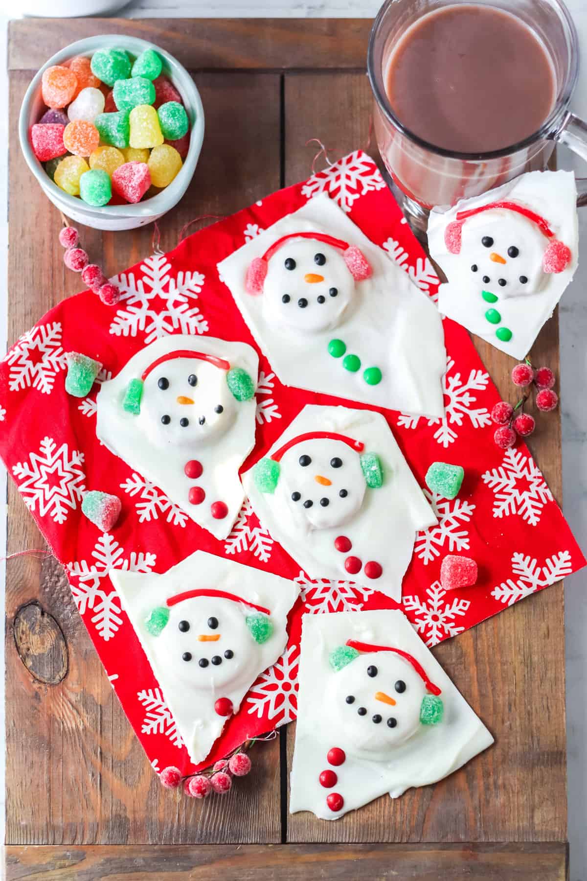 Individual pieces of finished melted snowman chocolate bark on serving tray with bowl of gumdrop candies and a glass of hot cocoa.